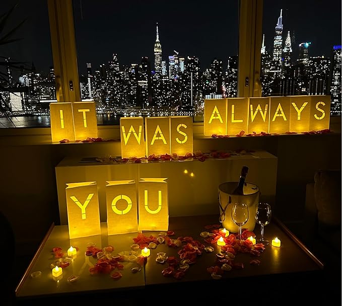 Romantic night setup with illuminated "It Was Always You" sign, rose petals, candles, wine, and a city skyline view